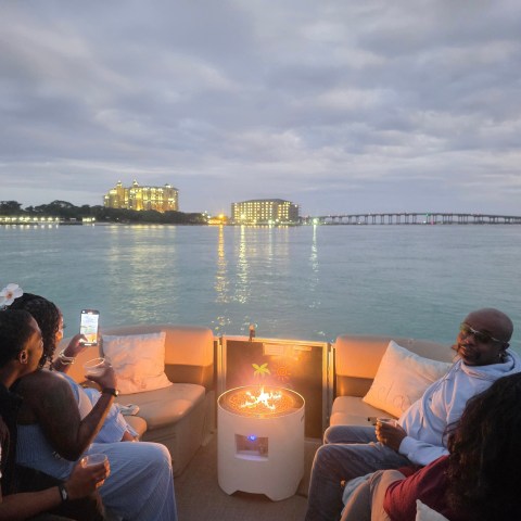 People on a boat with a fire pit, city skyline and a bridge in the background at dusk.