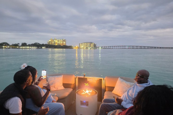 People on a boat with a fire pit, city skyline and a bridge in the background at dusk.