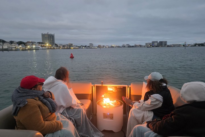 People on a boat with blankets around a fire pit on a cloudy day.