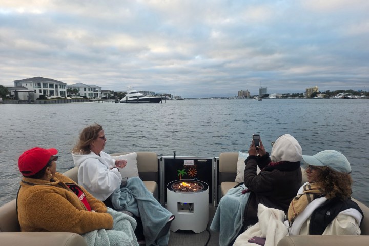 Four people sitting on a boat with blankets, near a fire pit, on a cloudy day, overlooking the water.