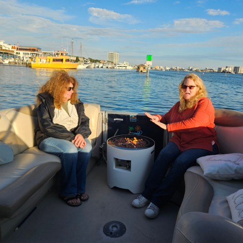 Two people seated on a boat deck with a small fire pit, surrounded by water and a city skyline in the background.