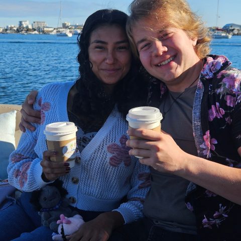 Two people sitting by water, smiling, holding coffee cups, wearing floral sweaters.