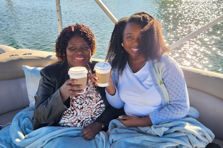 Two people on a boat with blankets, holding coffee cups, near water.