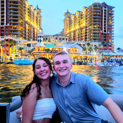 Two smiling people on a boat with illuminated buildings in the background.