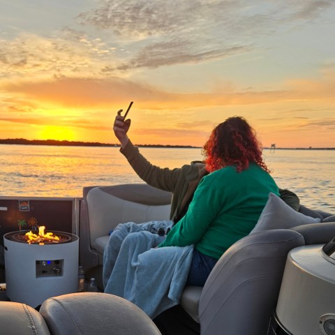 Two people on a boat at sunset, one holding a phone, with a small fire pit nearby.