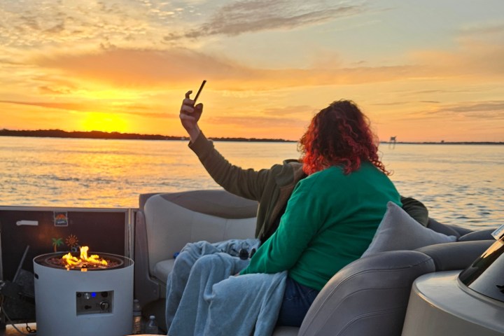 Two people on a boat at sunset, one holding a phone, with a small fire pit nearby.