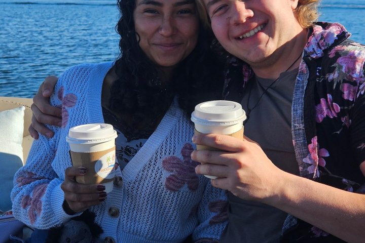 Two people smiling with coffee cups, sitting by a waterfront on a sunny day.