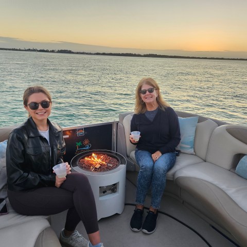 Two women on a boat with a fire pit, holding cups, at sunset over water.