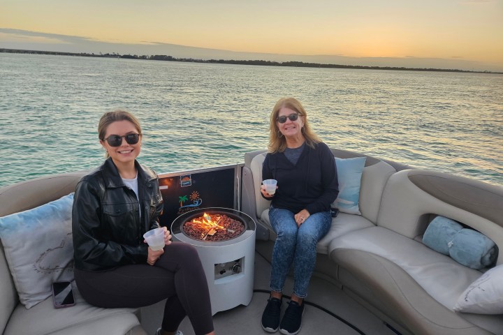 Two women on a boat with a fire pit, holding cups, at sunset over water.