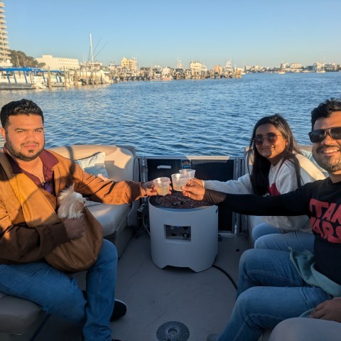Three people having drinks on a boat with a dog by a waterfront.