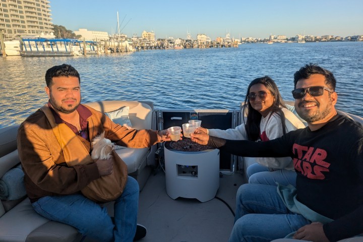 Three people having drinks on a boat with a dog by a waterfront.