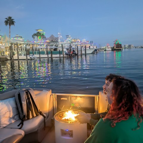 Two people on a boat with a central fire pit, colorful lights reflecting on the water at dusk.