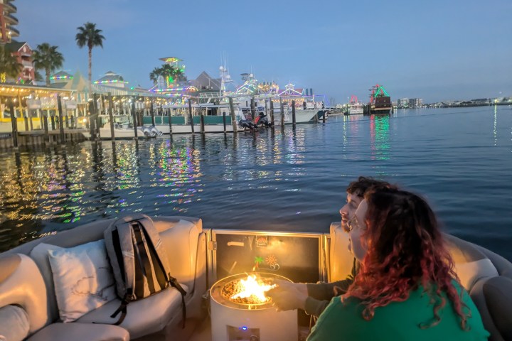Two people on a boat with a central fire pit, colorful lights reflecting on the water at dusk.