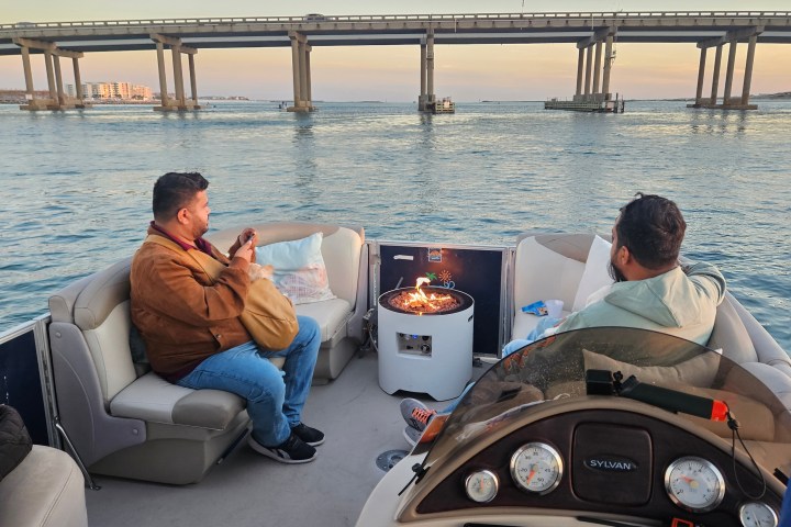 Two people on a boat with a small fire pit near a bridge at sunset.