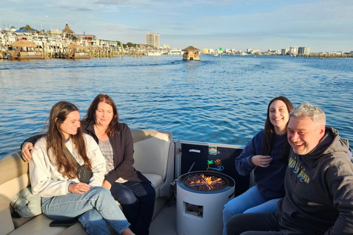 Four people seated on a boat with a fire pit, water and buildings in the background.