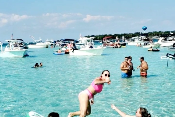 Person in pink swimsuit jumps off paddleboard into clear water with boats and people in background.