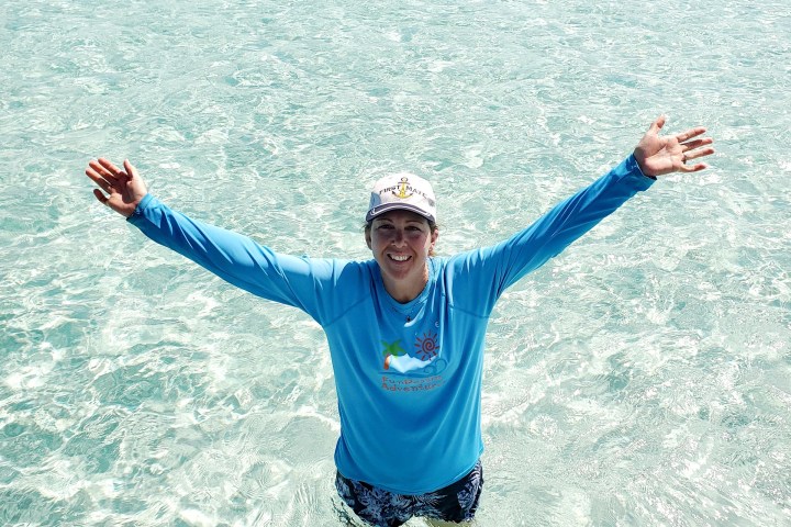 Person standing in clear shallow water, arms raised, wearing a blue shirt and white cap.
