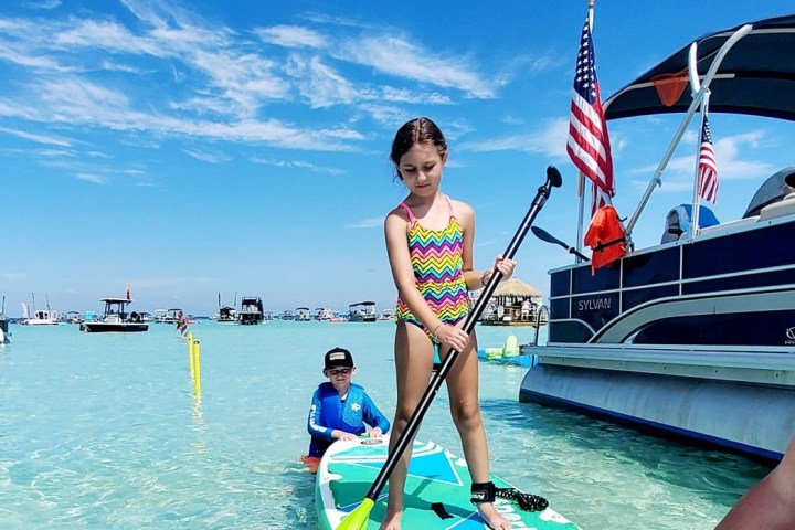 Child paddleboarding on clear water near a boat, with a blue sky.