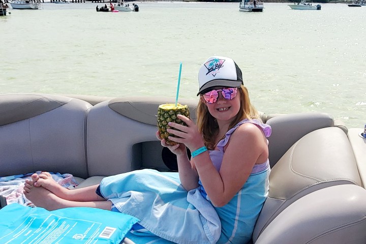 Girl on boat holding pineapple drink, wearing sunglasses and cap.