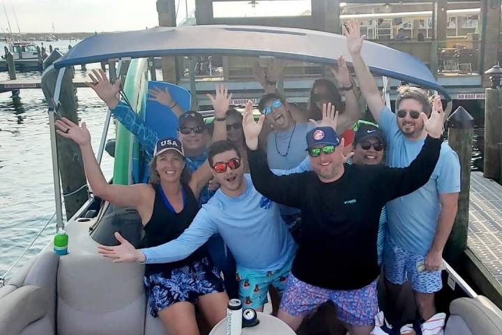 Group of smiling people on a boat, wearing casual summer clothes, with arms raised in excitement.