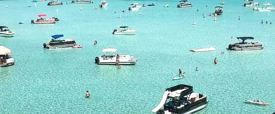 Boats and people on clear blue water near a bridge with a city skyline.