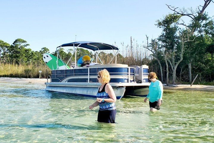 Two people wading in shallow water near a pontoon boat on a sunny day.