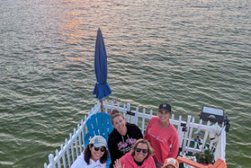 Group of five people on a deck by the sea at sunset, with chairs and fence visible.