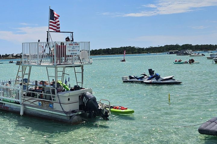 Pontoon boat with slide, docked in clear water, American flag on top, surrounded by jet skis and other boats.