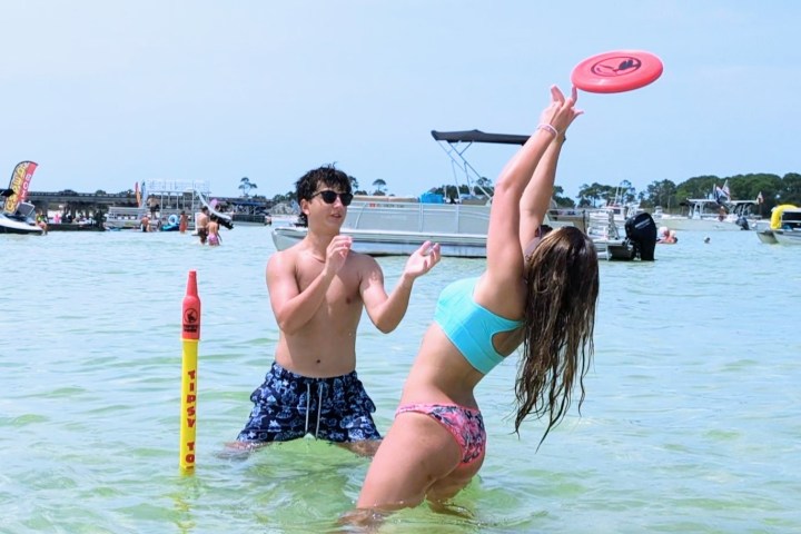 Two people playing catch with a frisbee in shallow water near boats.