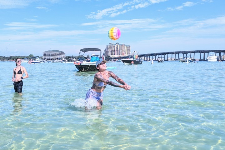 Person playing with a ball in shallow water near boats and a bridge.