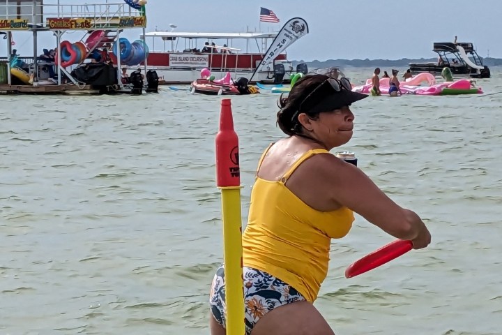 Person in yellow swimsuit plays frisbee near a lake with boats and floats in the background.