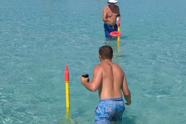 Two men playing frisbee in shallow water with boats in the background.
