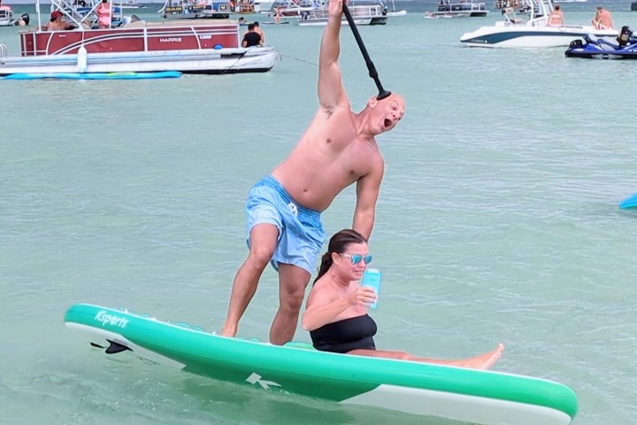 Man playfully posing on paddleboard with woman holding a drink, surrounded by boats on a sunny day.