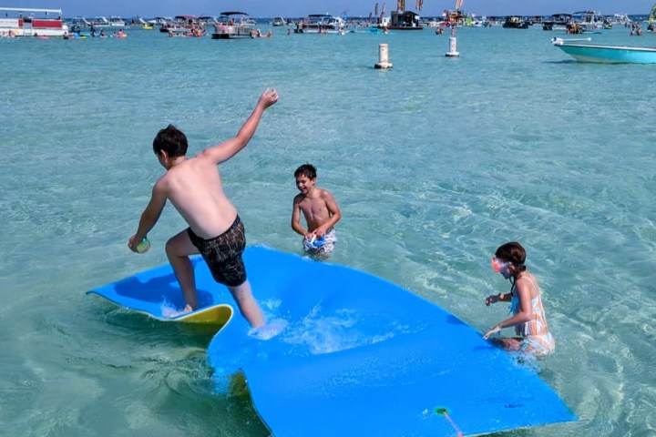 Kids playing on a floating mat in shallow water with boats in the background.