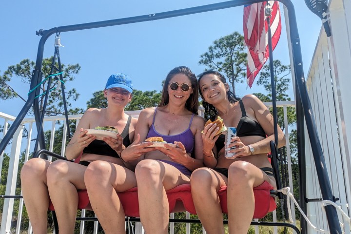 Three people on a swing seat, holding food and drinks, with an American flag in the background.
