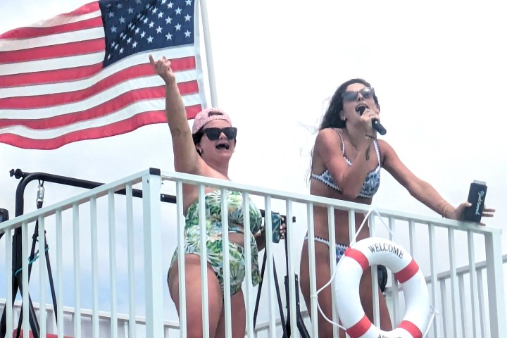 Two women on a boat balcony, one holding a microphone, the other with an American flag.