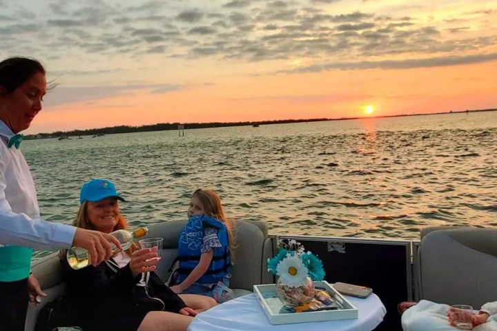 Server pours drinks on a boat at sunset with three people seated around a table.