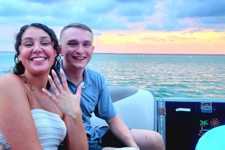 Smiling couple on a boat, woman showing a ring, ocean and colorful sunset in the background.