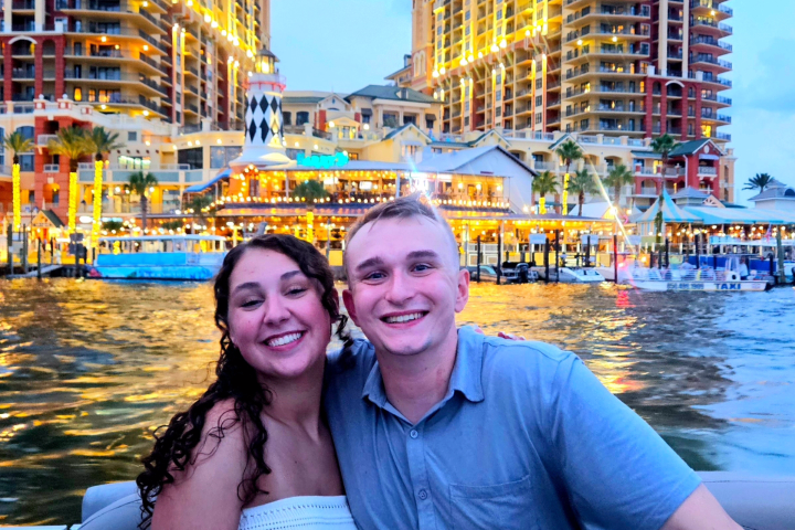 Smiling couple on a boat with lit buildings in the background at dusk.
