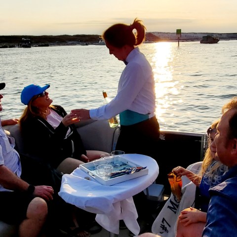People relaxing on a boat with drinks at sunset, served by a staff member.