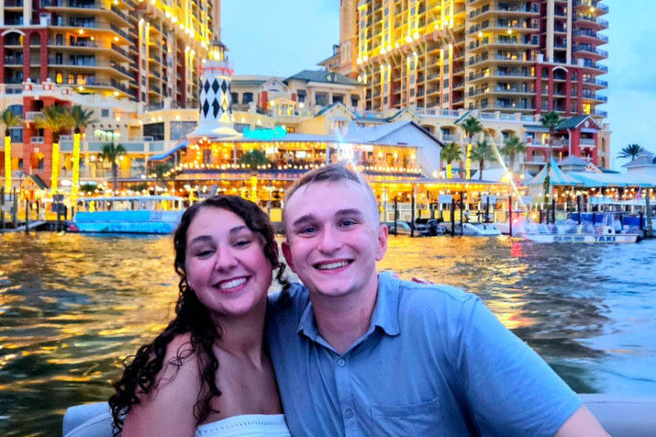 Smiling couple on a boat with illuminated buildings in the background.