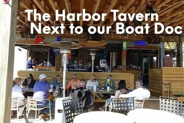 Outdoor bar with people sitting under a wooden roof, sign reads 'The Harbor Tavern Next to our Boat Doc'.